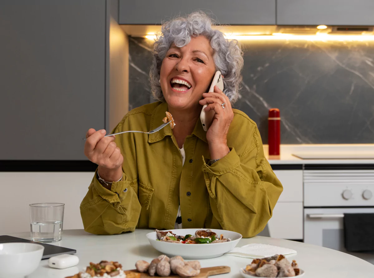 Smiling person talking on phone while eating at a kitchen table.