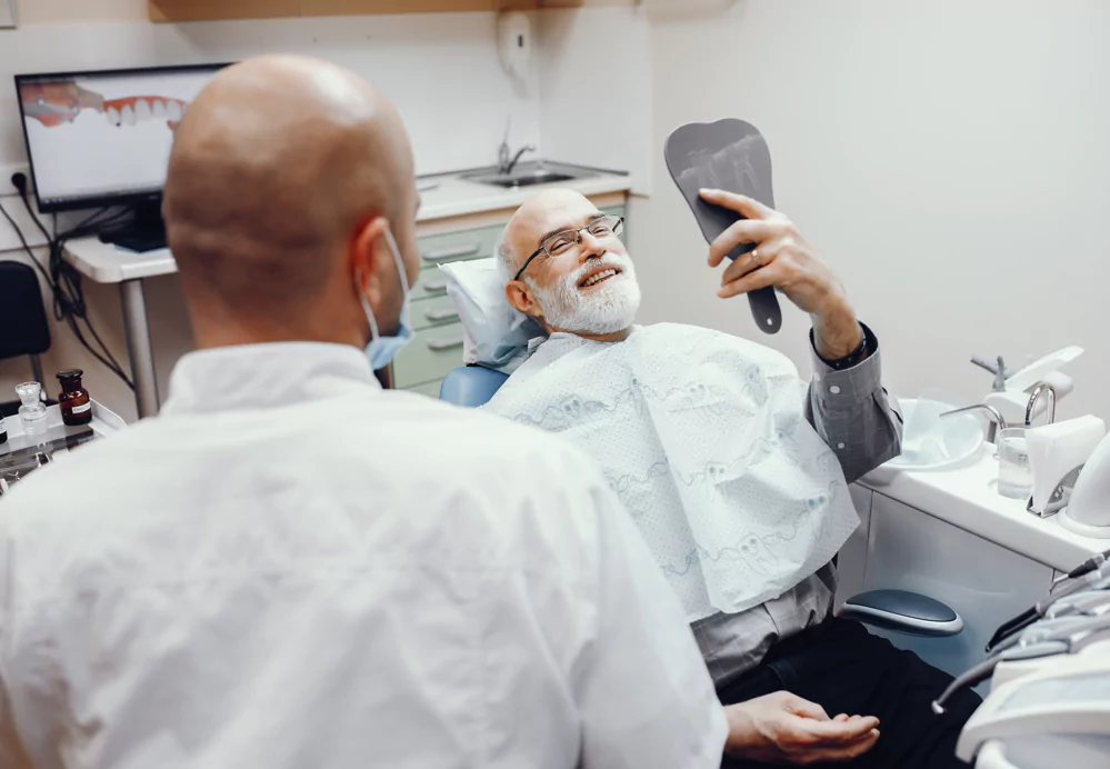 Elderly man smiling in dental chair, holding a mirror, with dentist nearby.