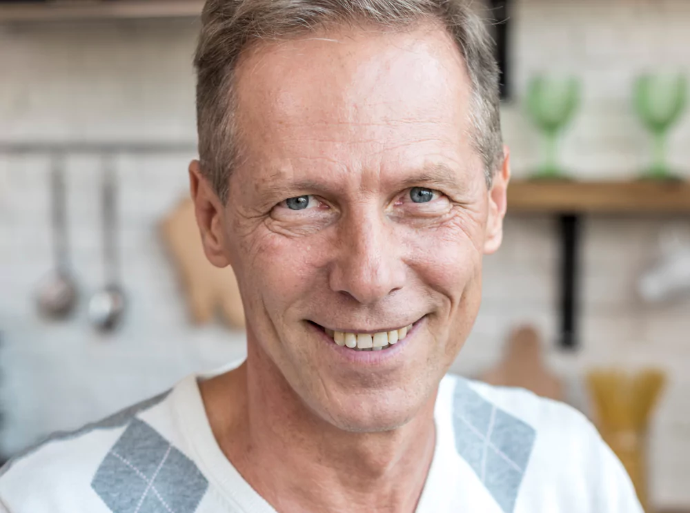 Smiling man in a kitchen with utensils in the background.