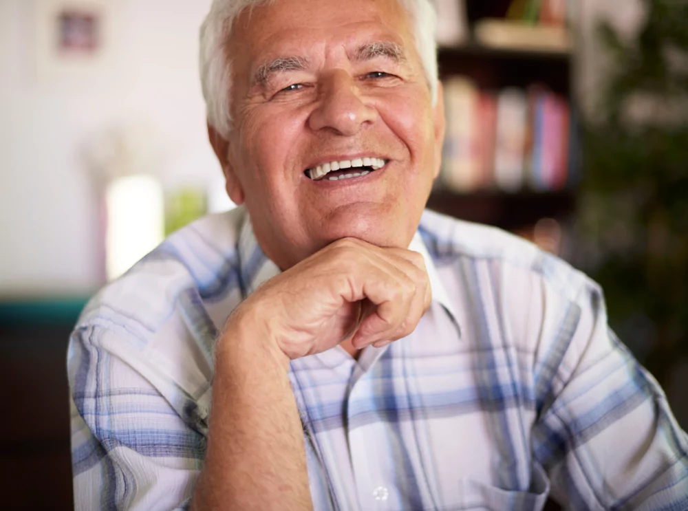 Elderly man smiling, wearing a plaid shirt, with hand under chin.