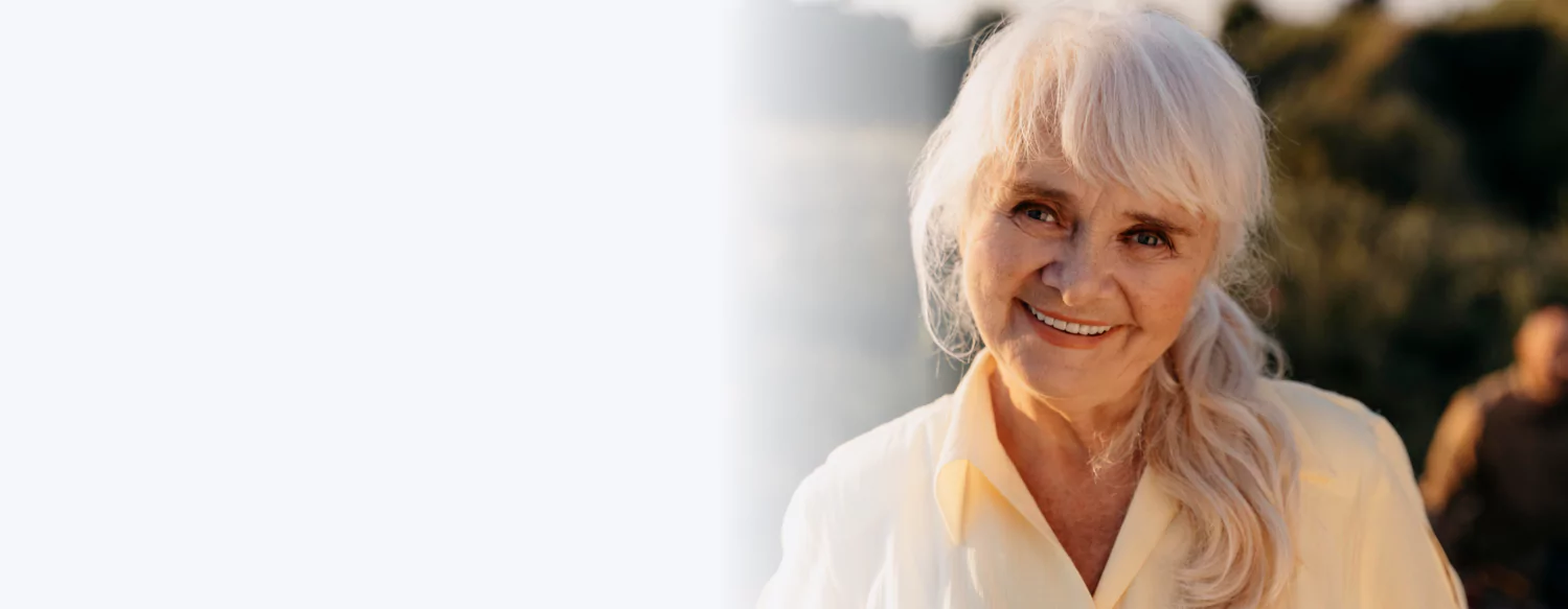 Smiling elderly woman with white hair in a yellow shirt outdoors.