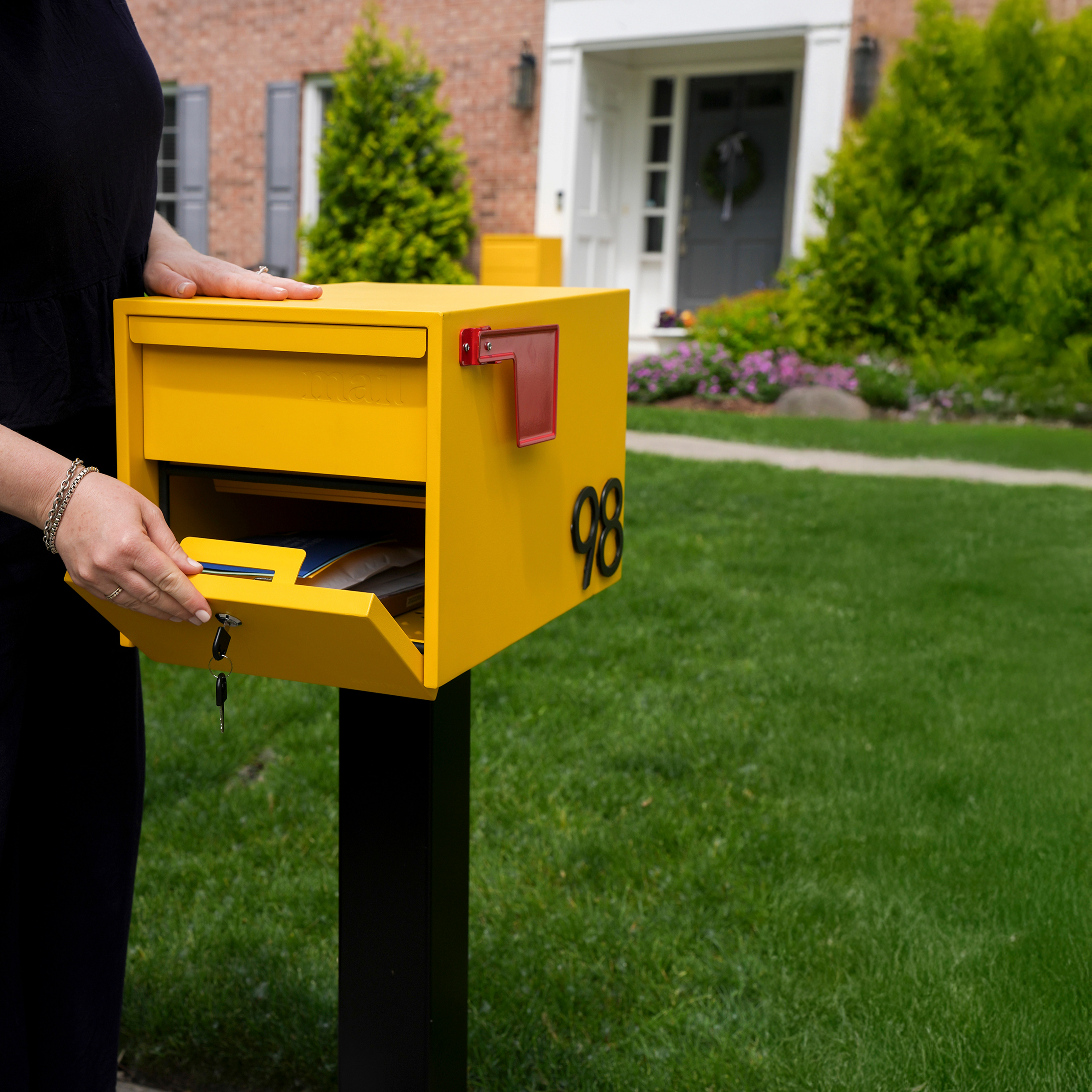Person opening a yellow mailbox with number 98 in a garden.