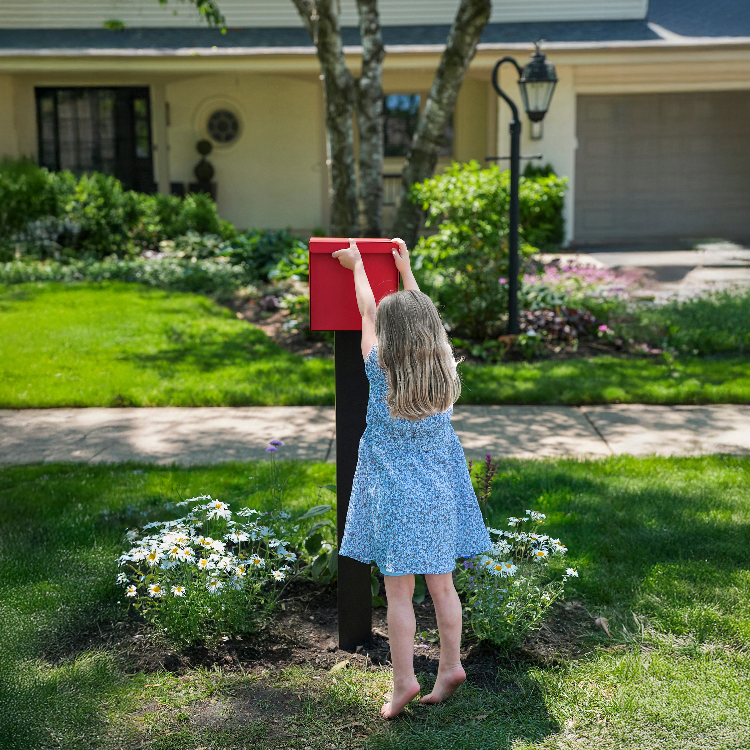 Young girl in blue dress reaching for a red mailbox in a garden.