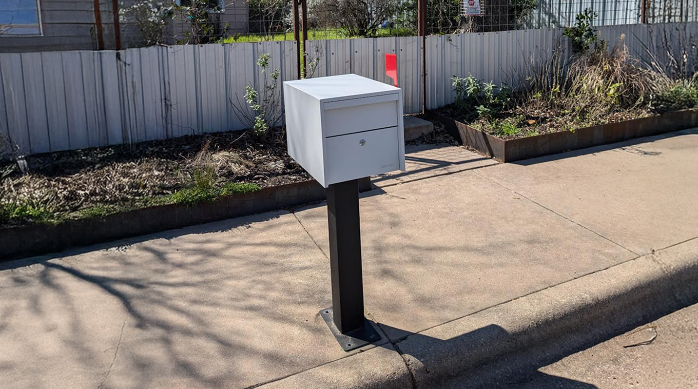 White mailbox on a black post by a sidewalk, with a fence and plants in the background.