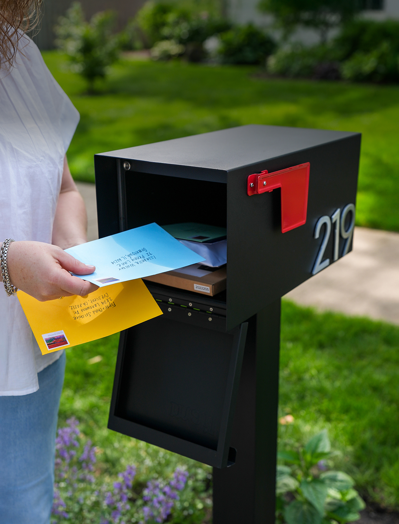 Person placing colorful envelopes into a black mailbox with a red flag.