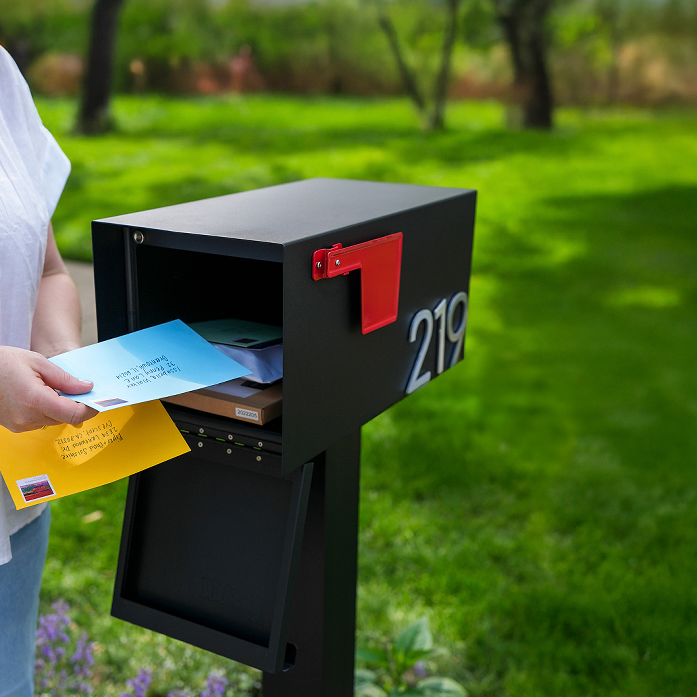 Person placing colorful envelopes into a black mailbox with red flag.