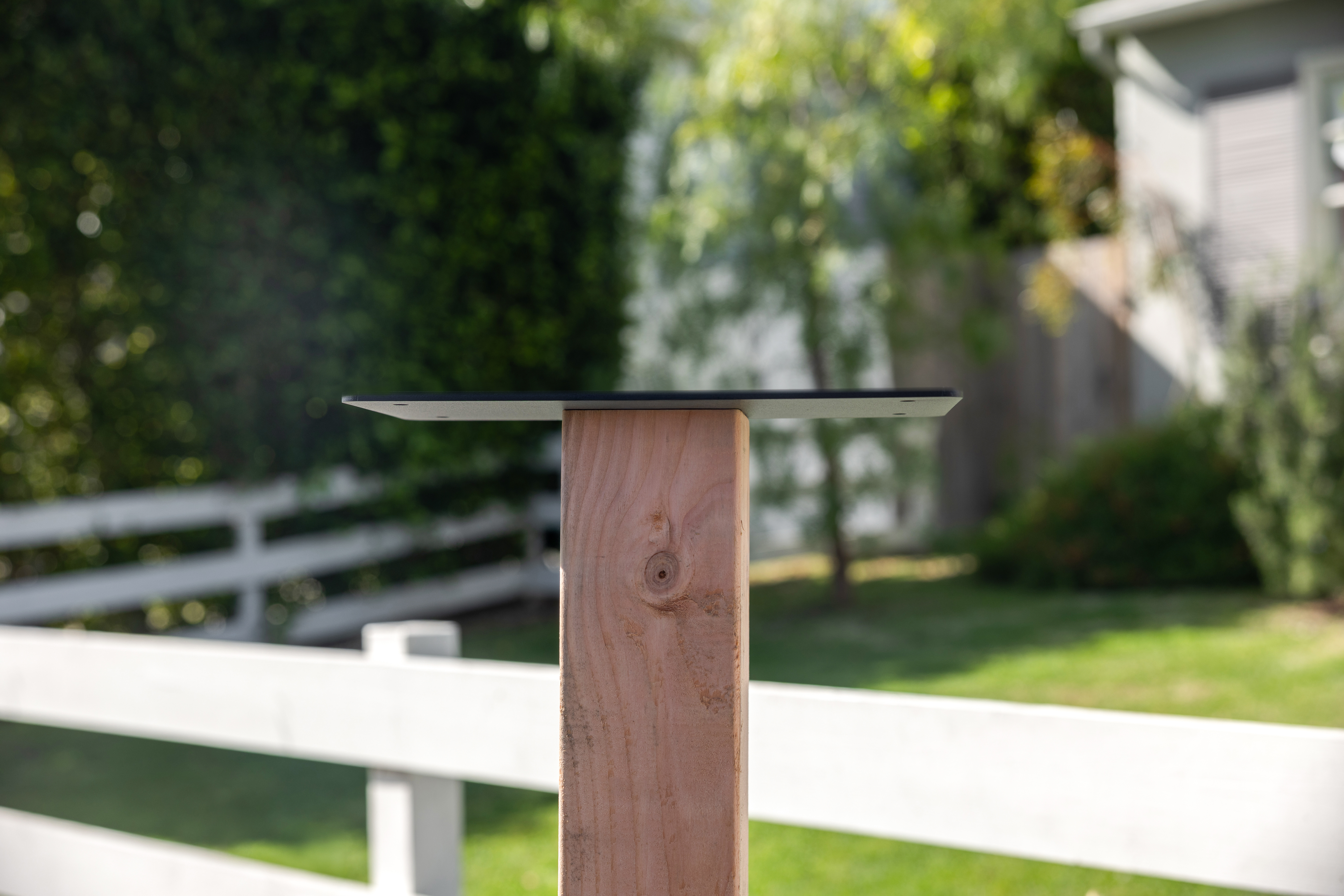 A black metal top plate mounted on a wooden post outdoors in front of a white fence.