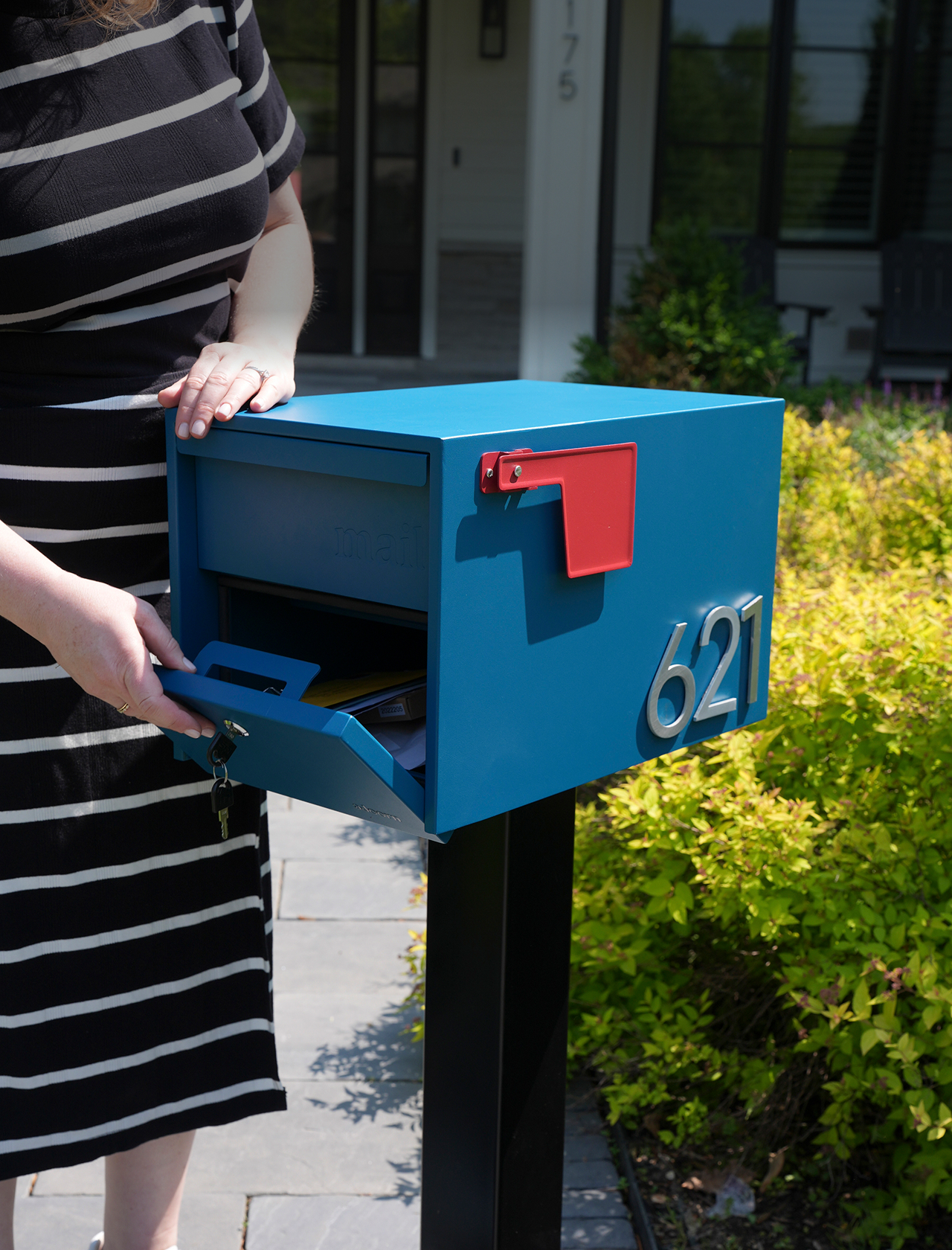Person opening a blue mailbox with the number 621, surrounded by greenery.
