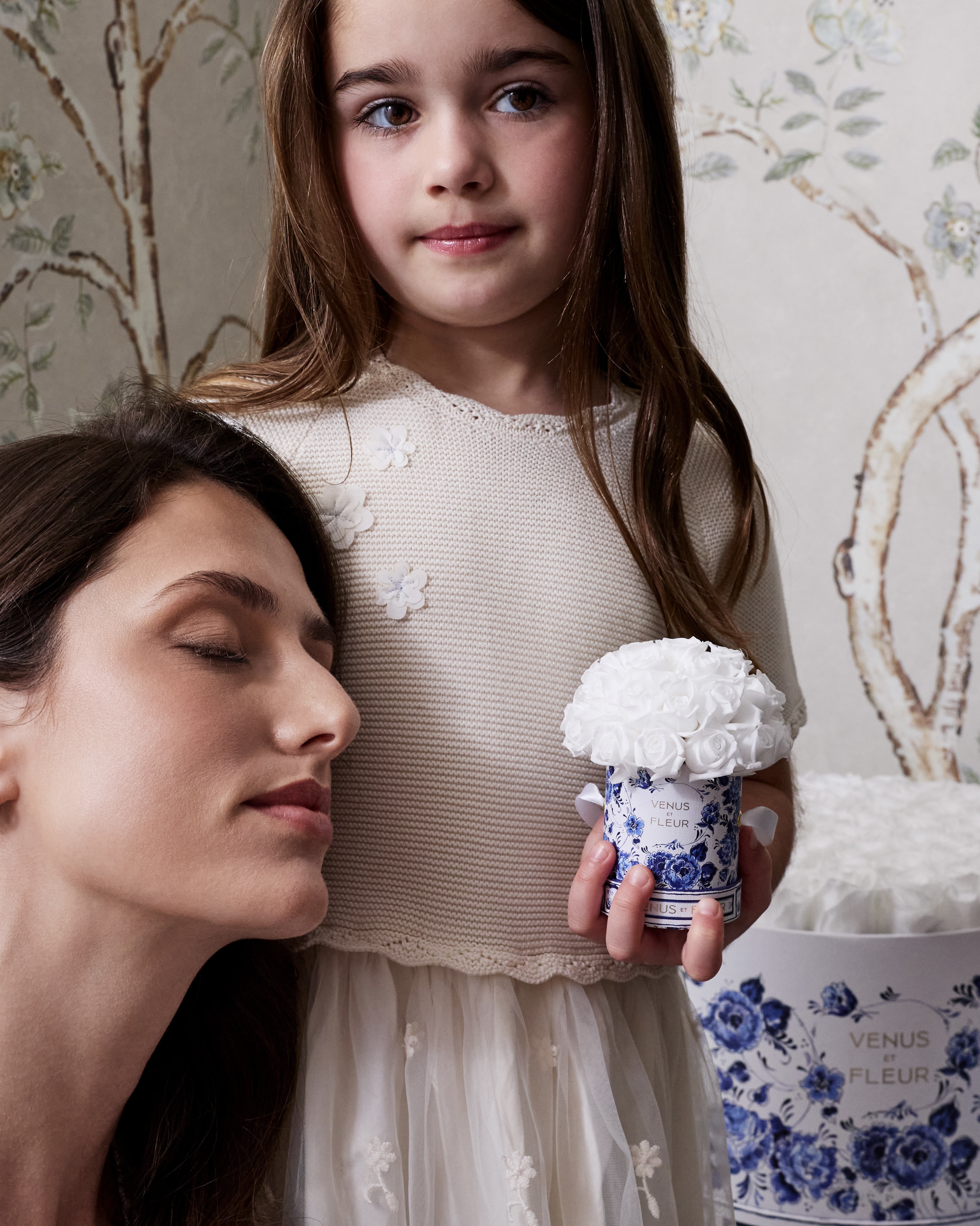 A young girl stands behind a woman with her eyes closed, holding a small box of white roses.