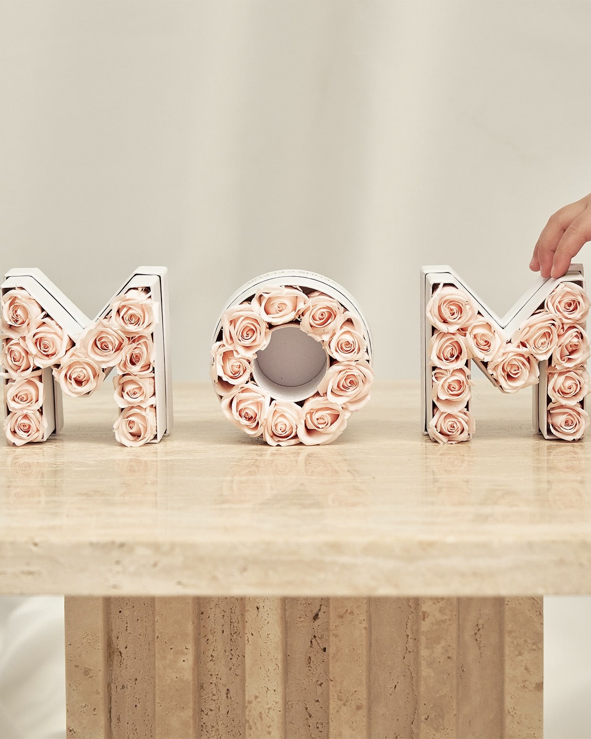 Letters spelling MOM filled with pink roses sit on a marble tabletop, with a hand touching one letter.