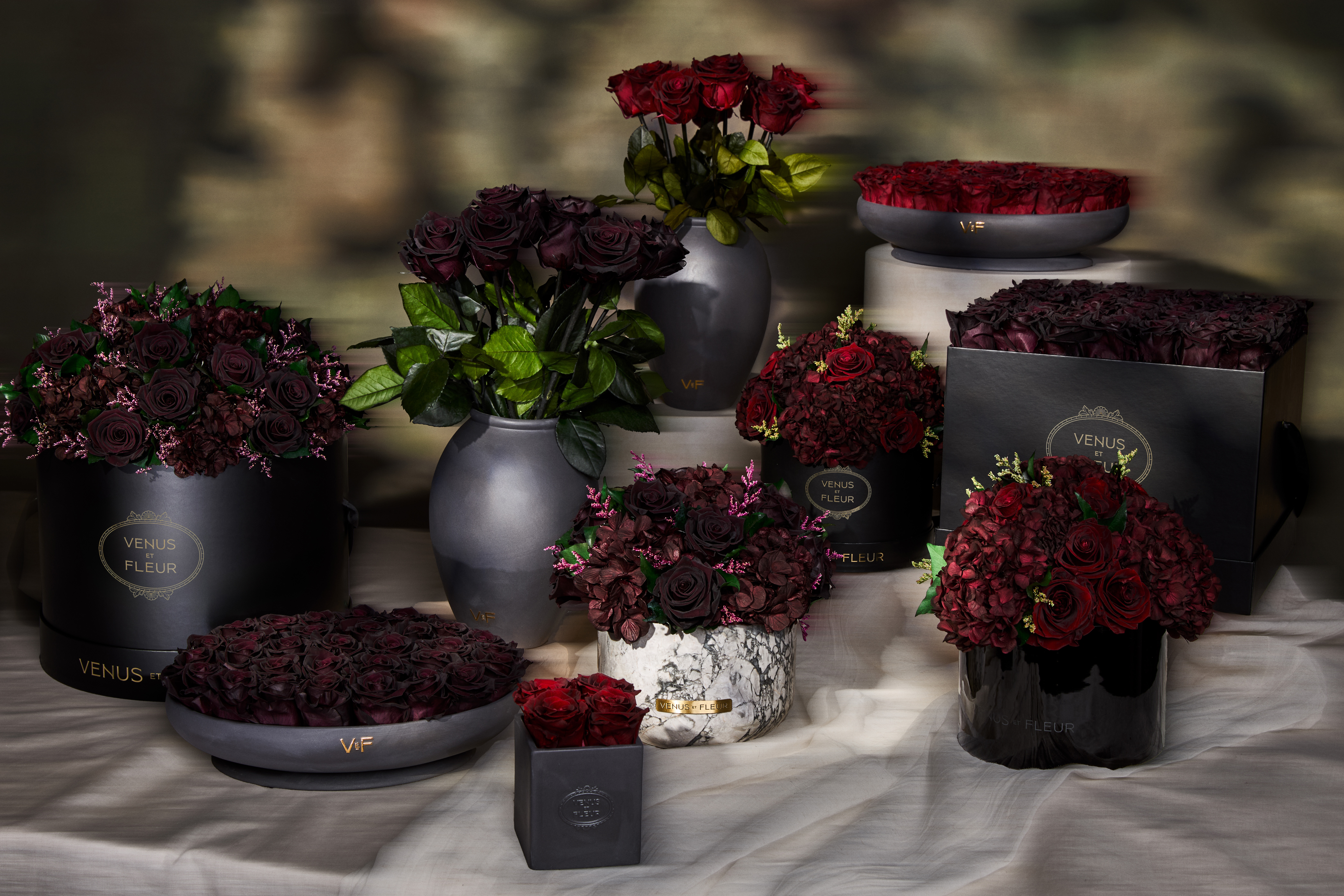 Assortment of dark red rose arrangements in various black vases and boxes.