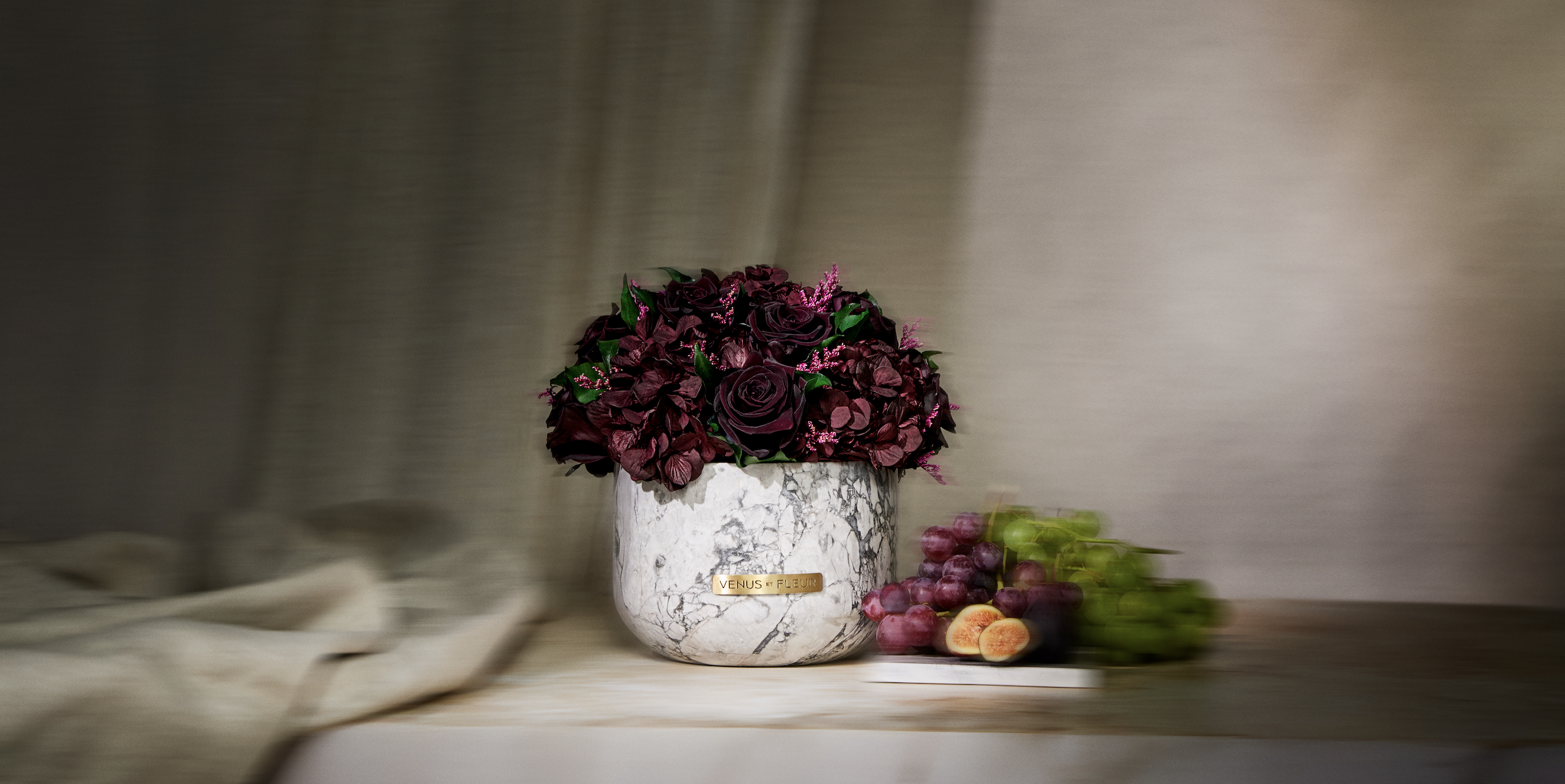 Marble vase with dark flowers, grapes, and figs on a table.
