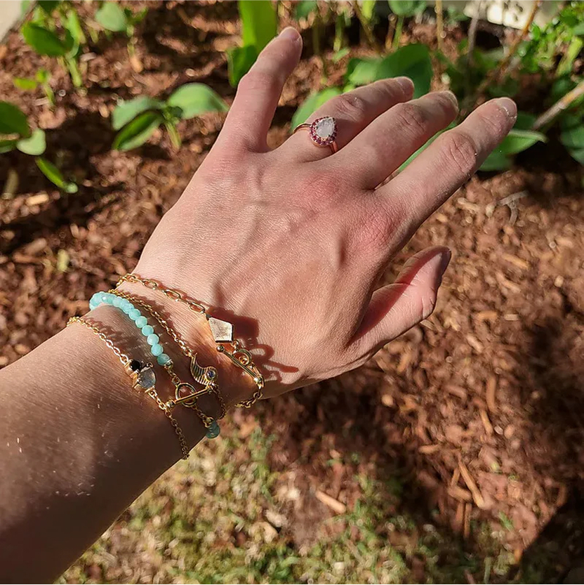 Hand with bracelets and a ring, outdoors with plants in the background.