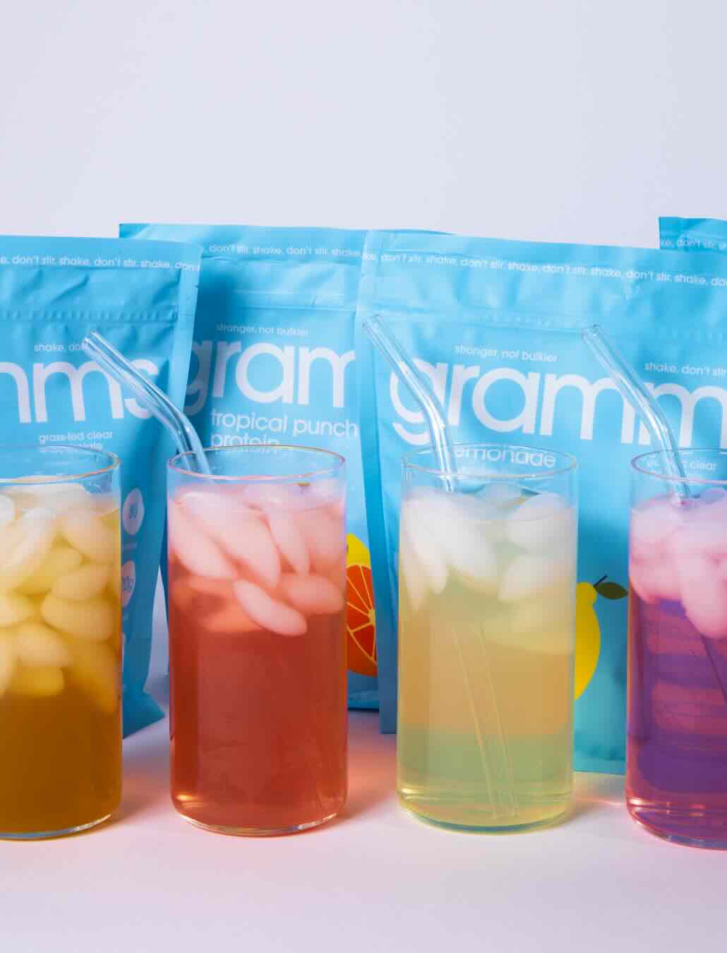 Four glasses of colorful iced drinks with straws stand in front of light blue product pouches.