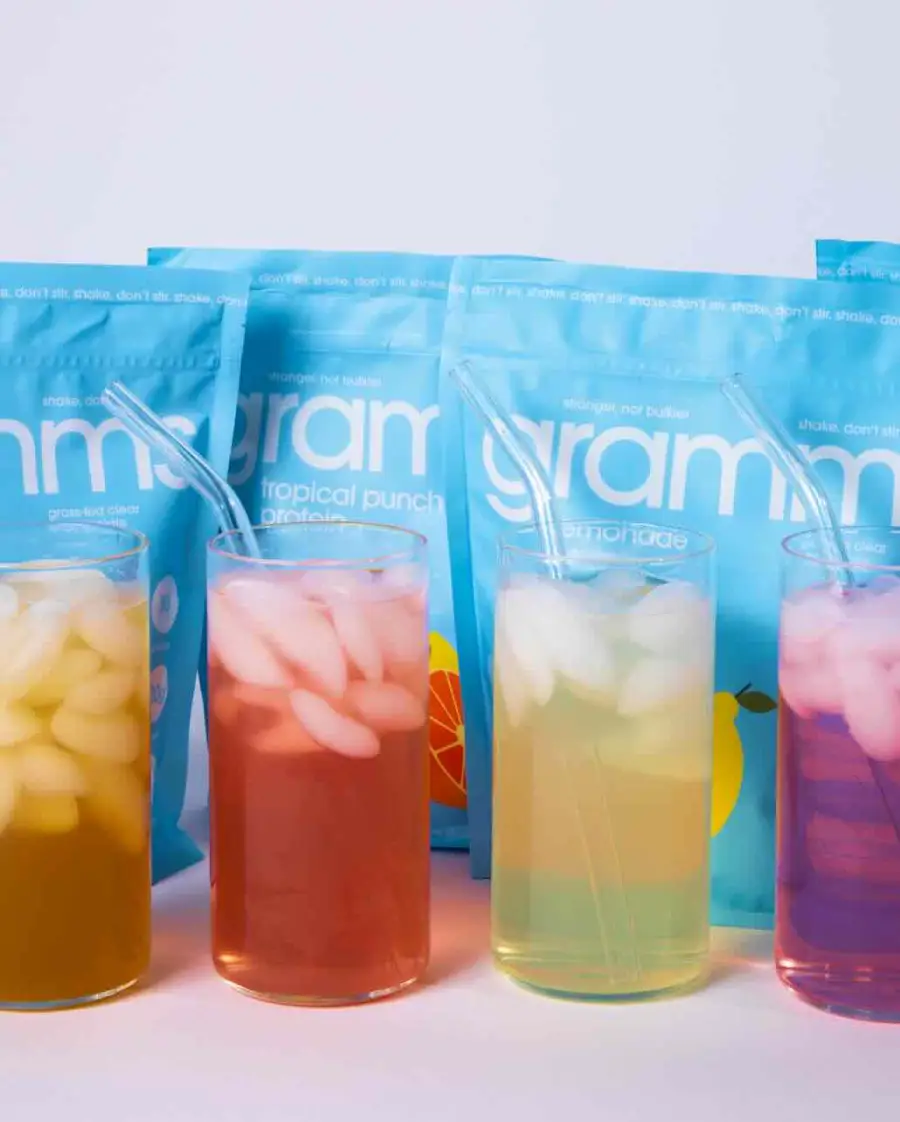 Four glasses of colorful iced drinks with glass straws sit in front of light blue product pouches.