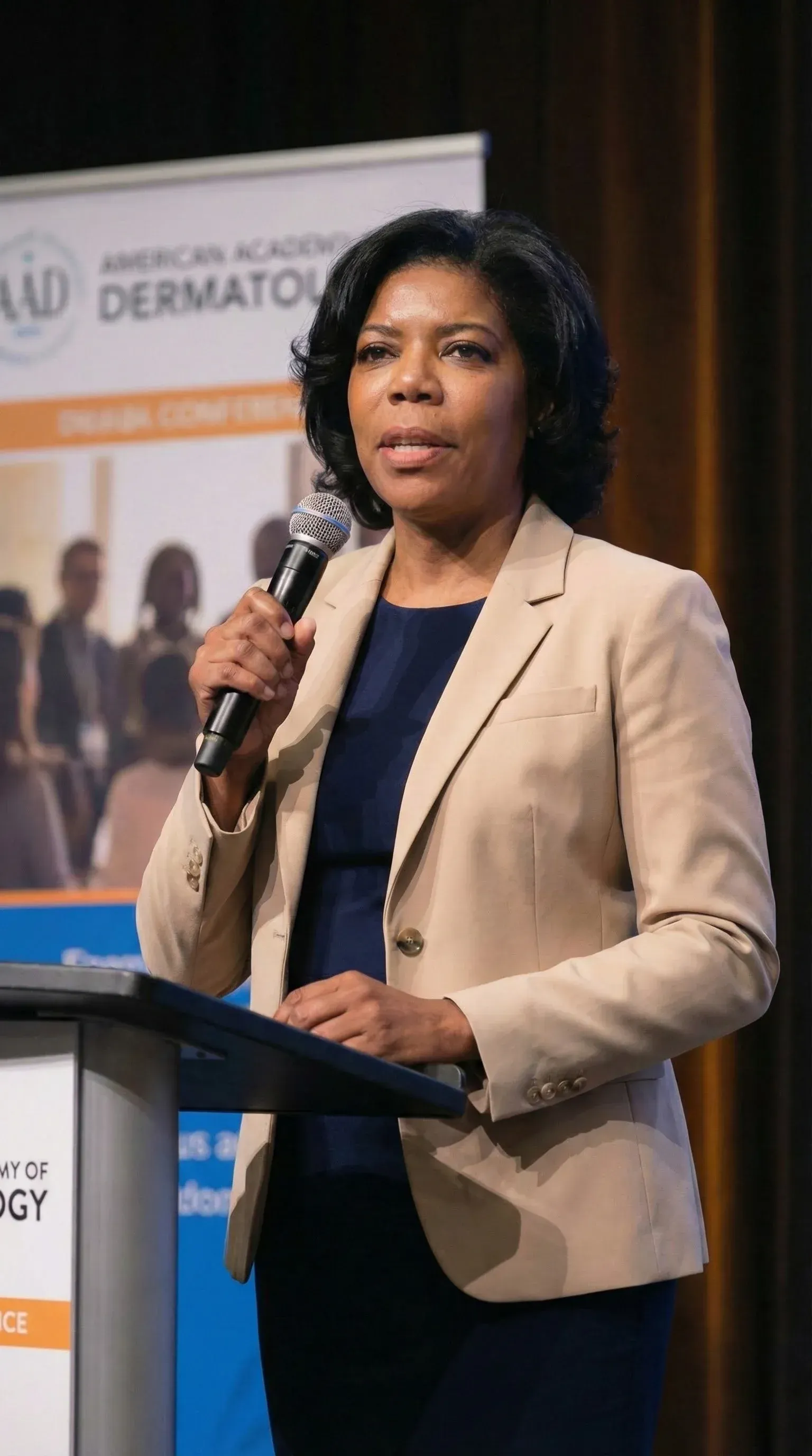 A woman in a beige blazer speaks into a microphone while standing at a podium during a conference.