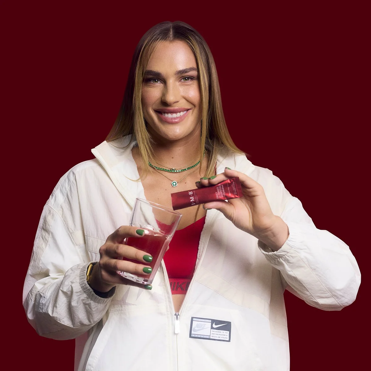 A woman in a white jacket smiles while squeezing a red supplement packet into a glass of liquid.
