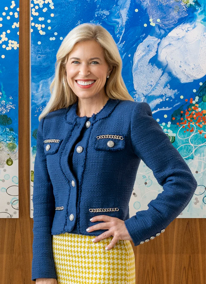 A smiling blonde woman in a blue tweed jacket and yellow houndstooth skirt, posing before abstract art.