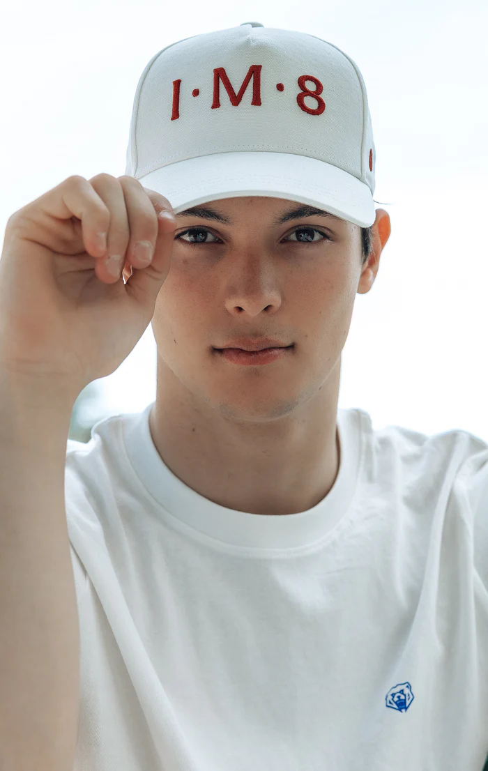 A young man in a white t-shirt and baseball cap looks at the camera, holding his cap's brim.