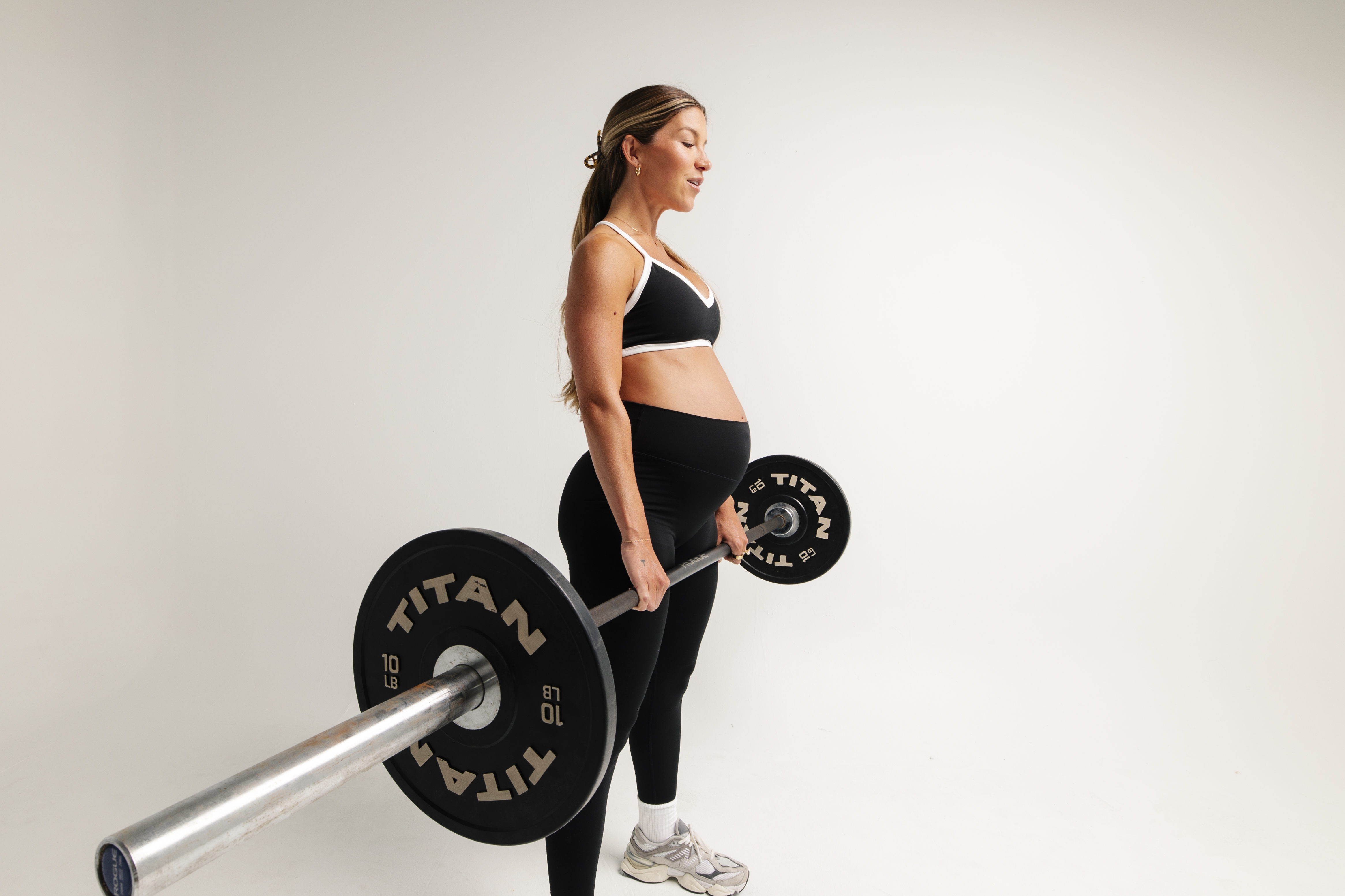 A pregnant woman in workout clothes stands holding a barbell with weights against a white background.