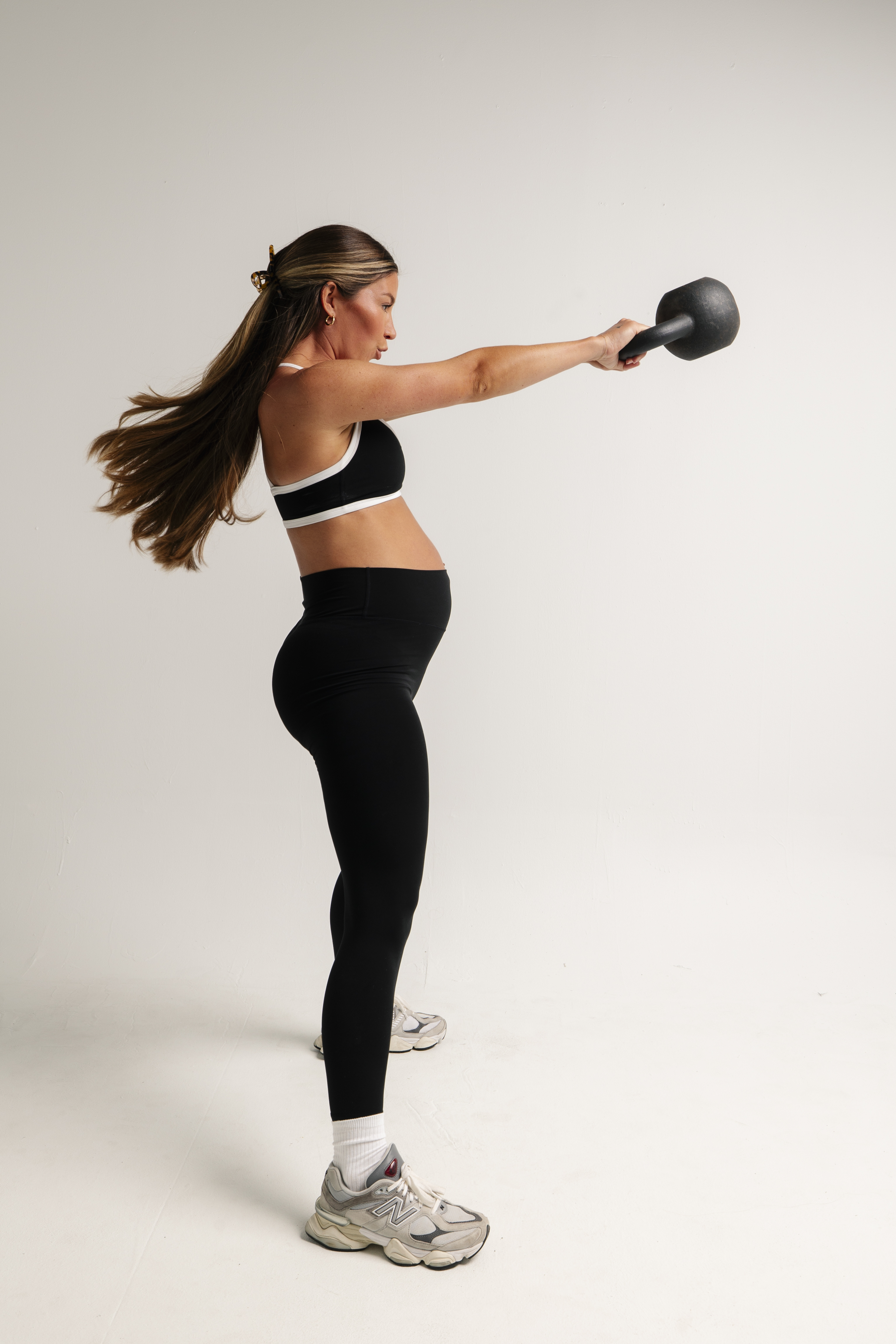 A pregnant woman in profile, wearing athletic clothes, swings a kettlebell against a white background.