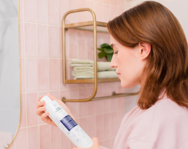 Person holding a bottle in a bathroom with pink tiles and a shelf.