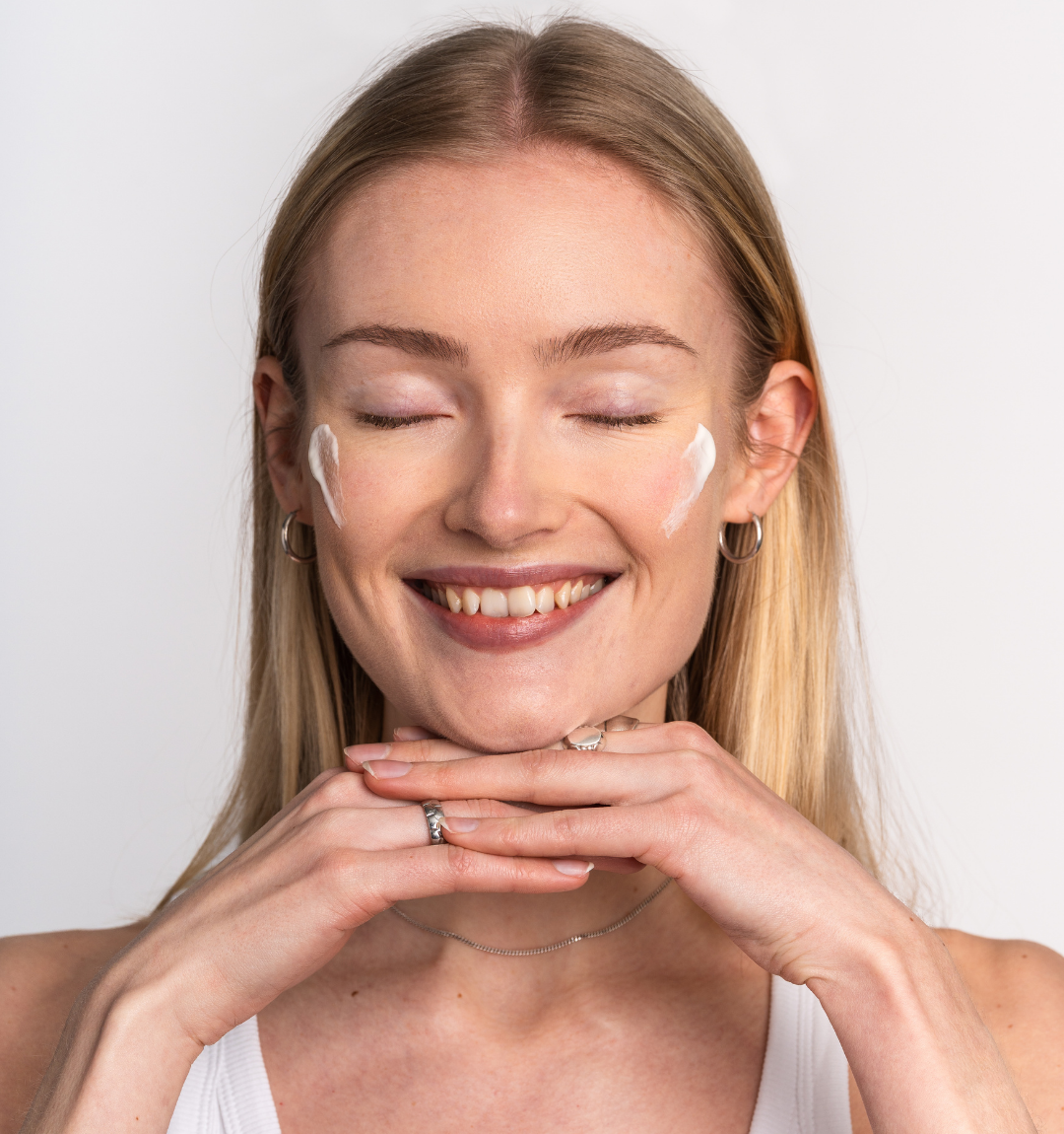 A smiling blonde woman with her eyes closed and dabs of white cream on her cheeks.