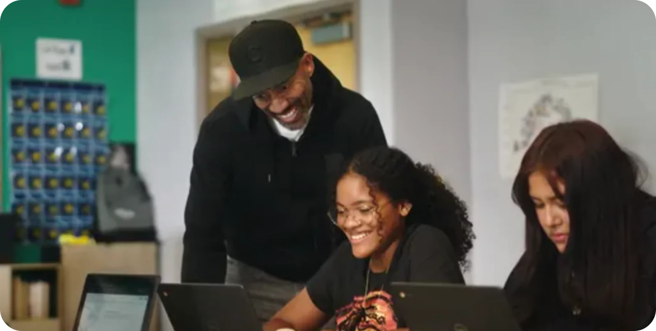 A teacher smiling at students working on laptops in a classroom.