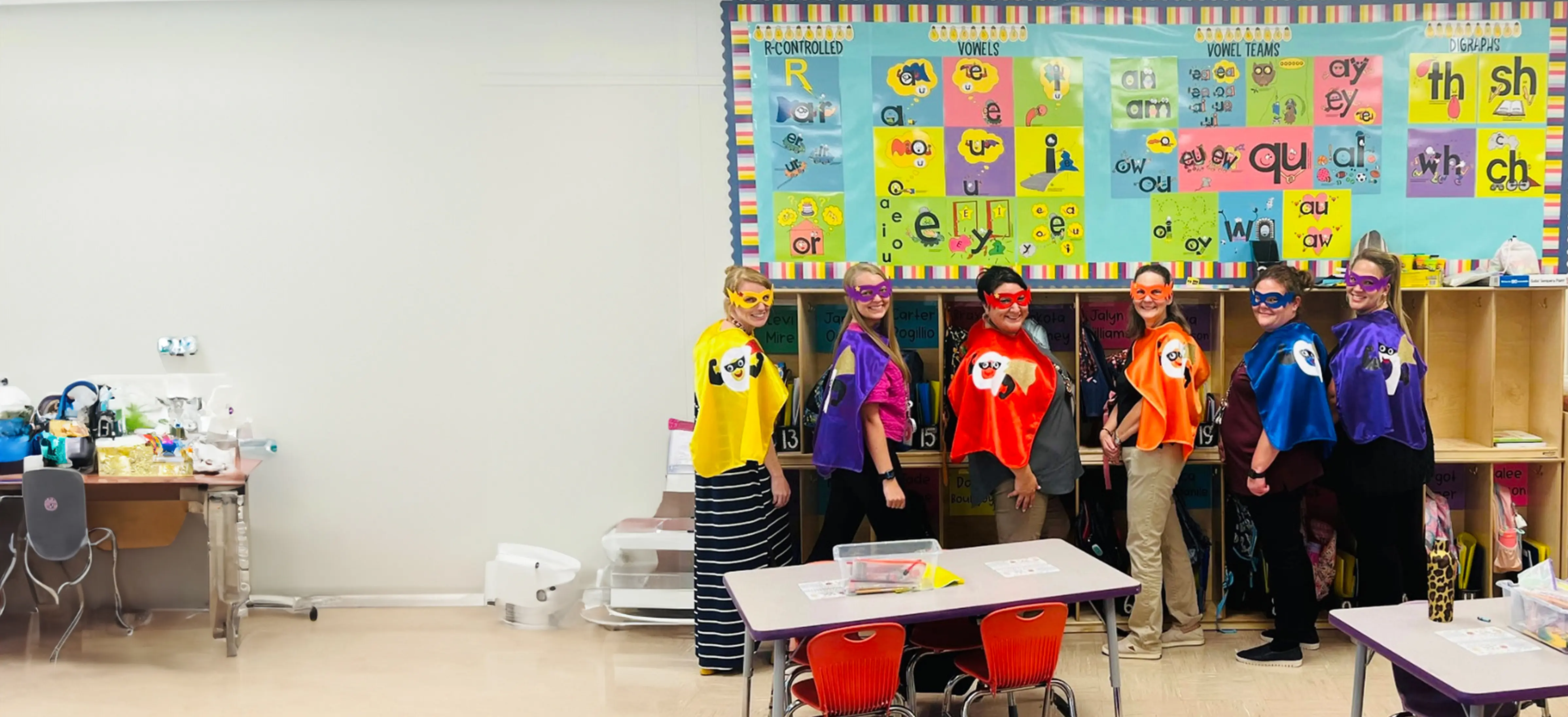 Six people in colorful superhero capes and masks in a classroom.