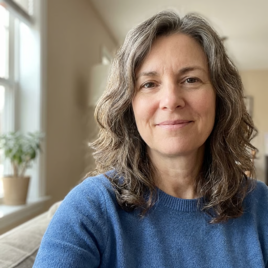 A headshot of a smiling woman with wavy, graying hair, wearing a blue sweater indoors.