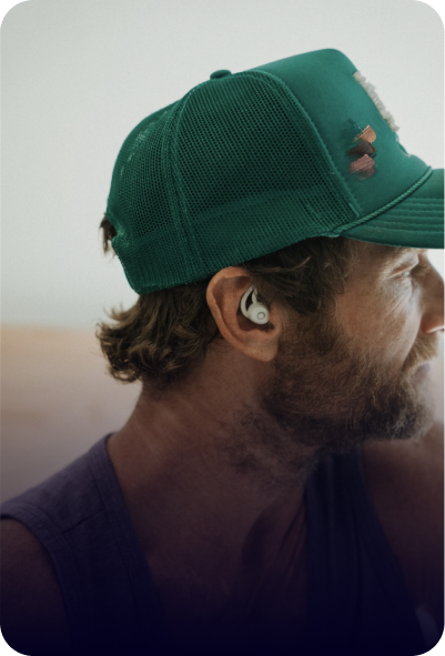 A man with a beard wearing a green trucker hat and a white earbud in his right ear.