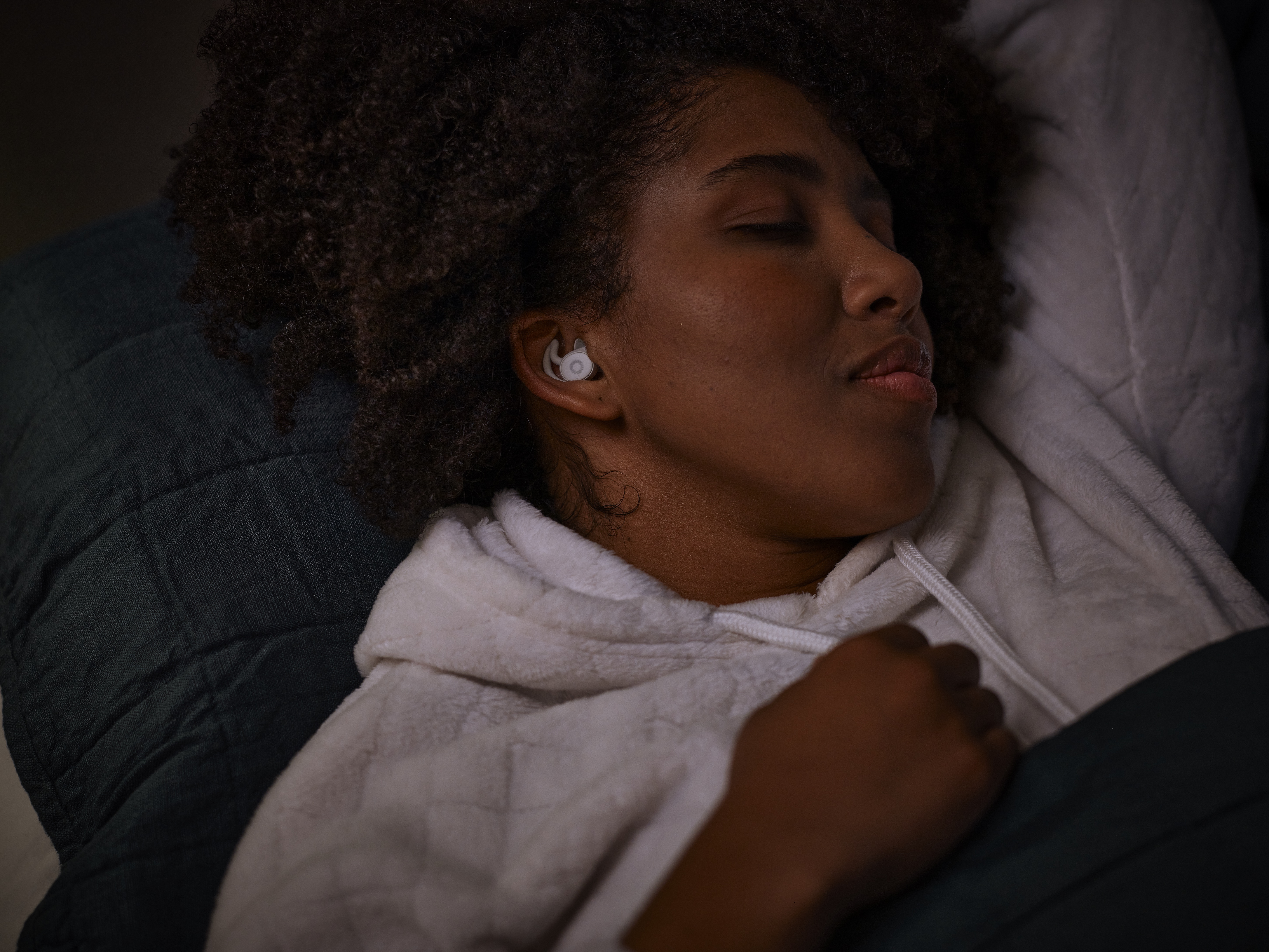 Person sleeping with earbuds, wearing a white hoodie, lying on a dark pillow.