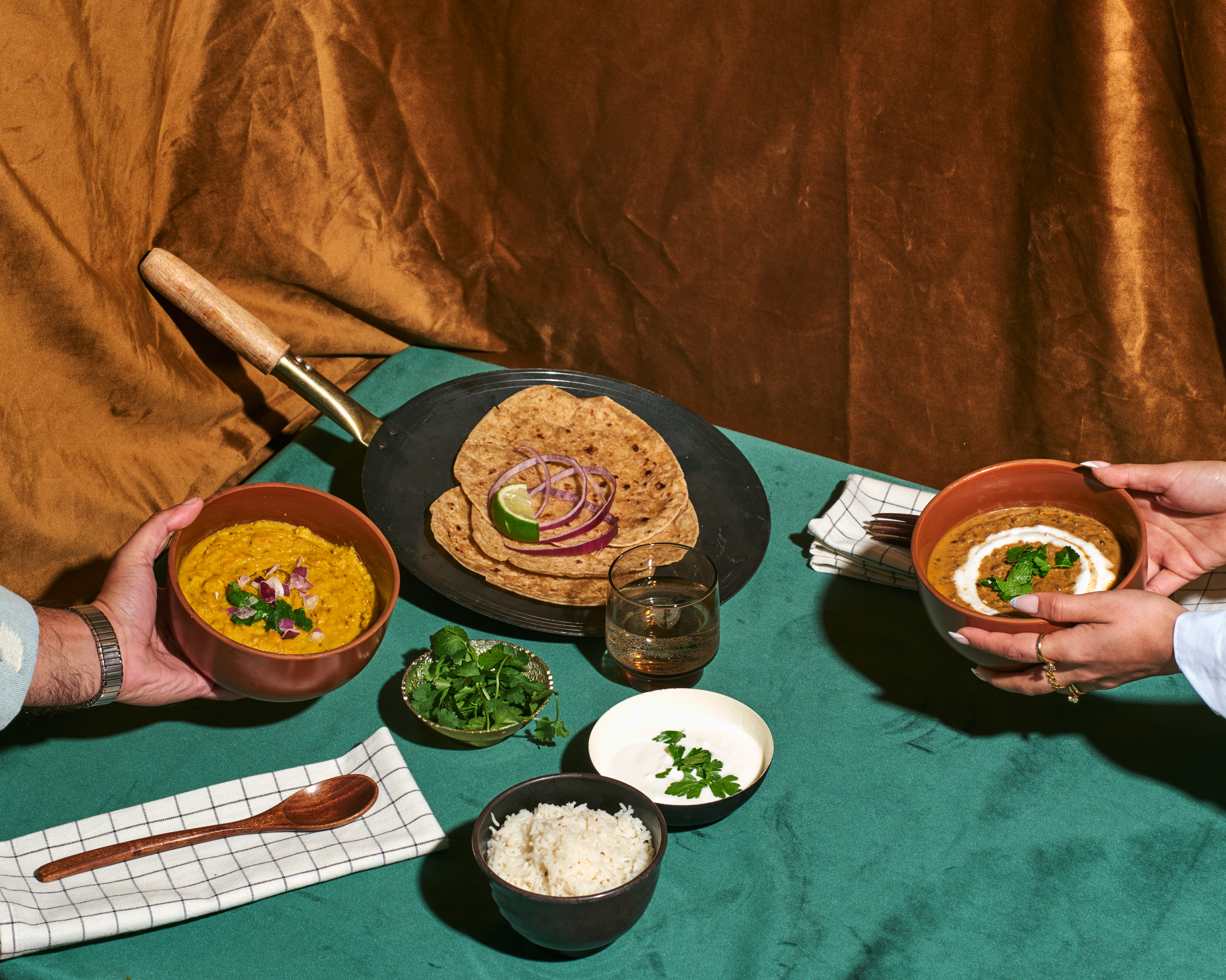 Two hands holding bowls of curry with naan, rice, and garnishes on a green tablecloth.