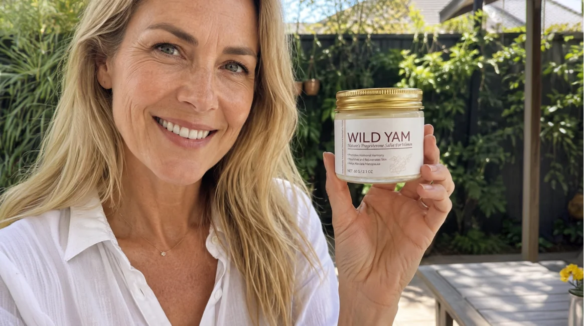 A smiling blonde woman in a white shirt holds up a jar of Wild Yam salve outdoors.