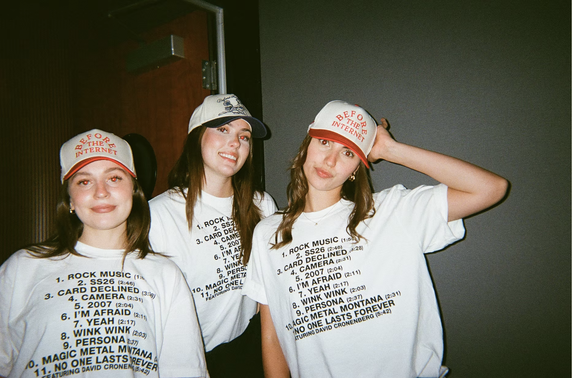 Three women in matching tracklist t-shirts and baseball caps pose for a film-style photograph.