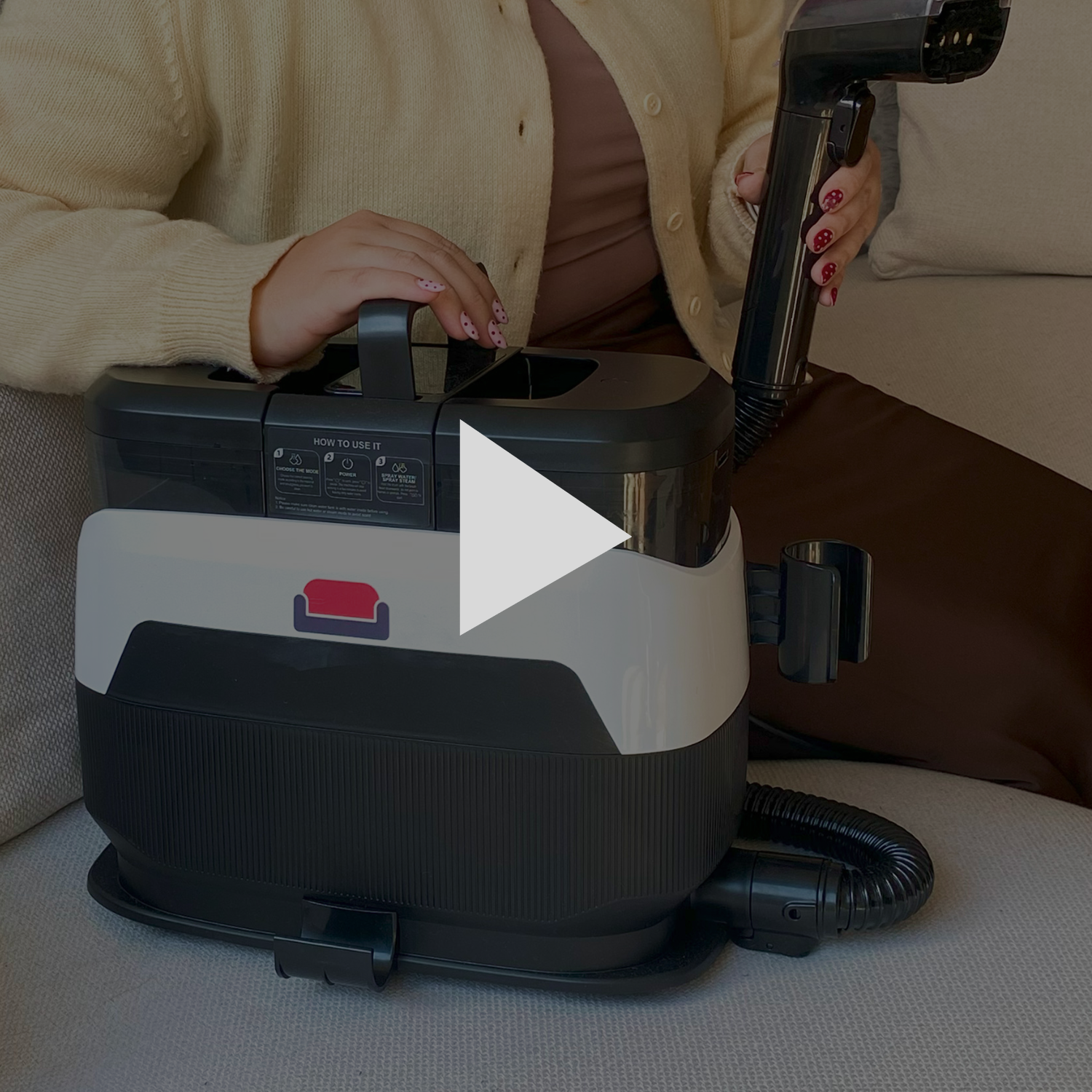 A person sitting on a couch holds the hose of a portable upholstery cleaner, with a play button overlay.