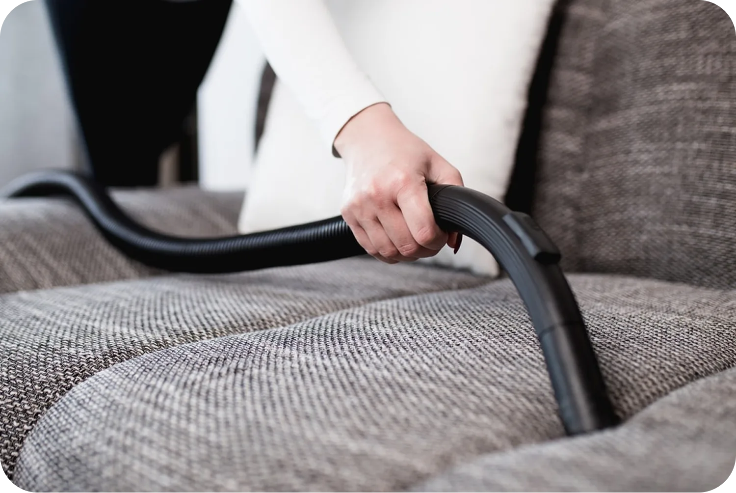 A person uses a vacuum cleaner hose attachment to clean a gray fabric sofa.