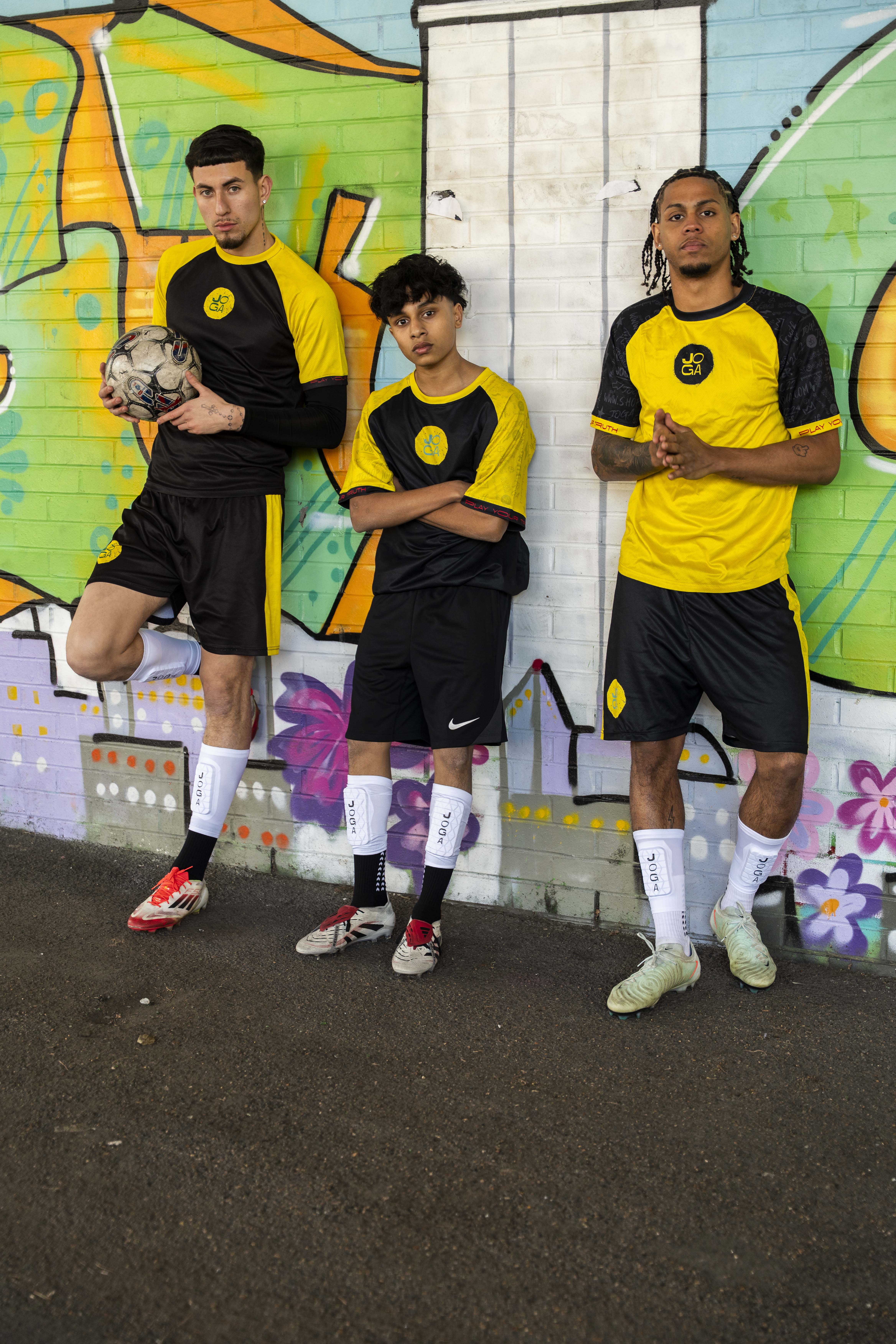 Three soccer players in yellow and black uniforms pose against a graffiti wall.