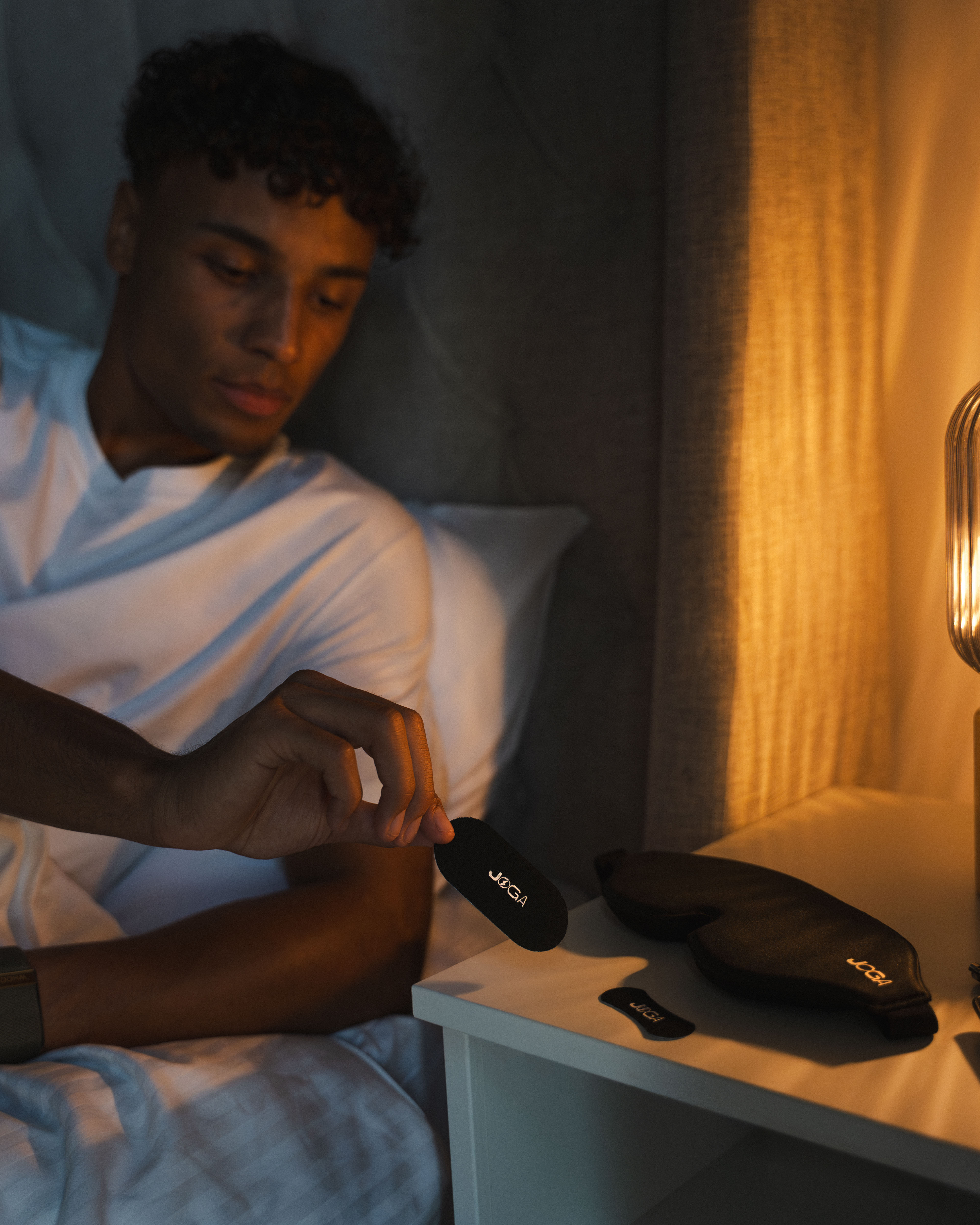 Person in bed holding a small black item near a nightstand with a sleep mask.