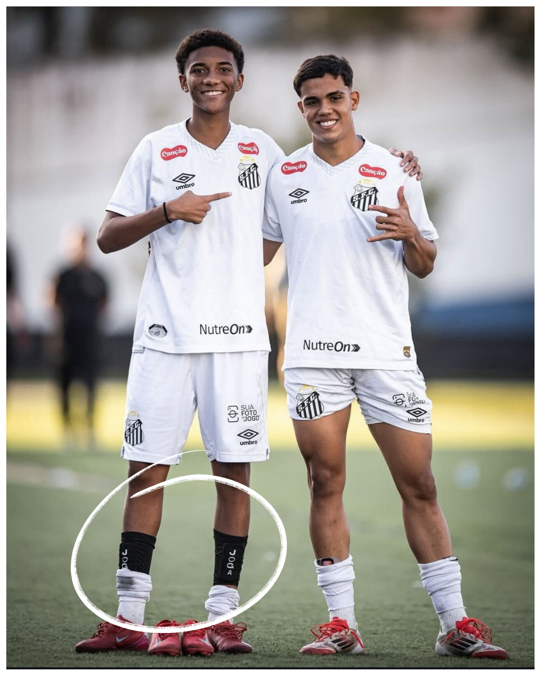 Two soccer players in white uniforms smiling and pointing at each other on a field.