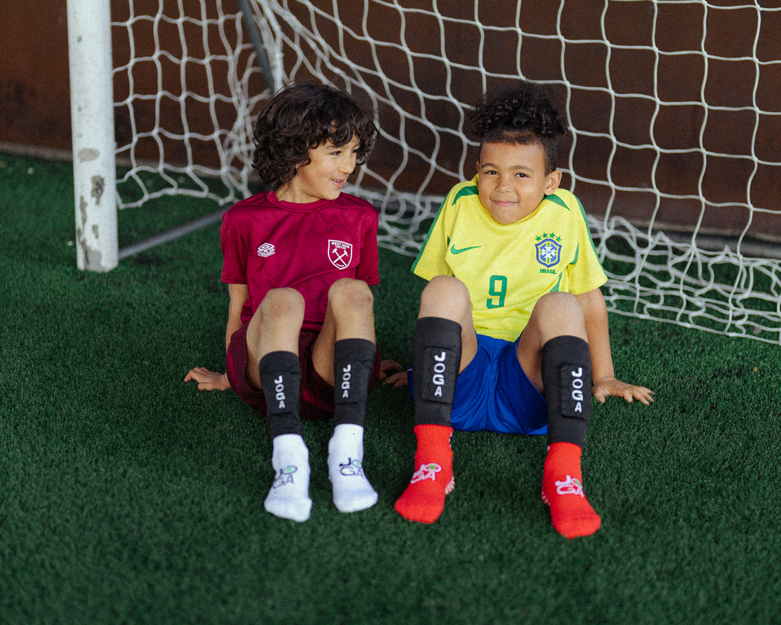 Two children in soccer uniforms sitting on grass in front of a goal net.