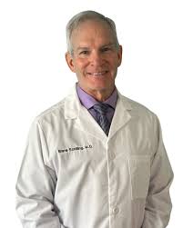 A smiling man with graying hair wearing a white doctor's coat over a shirt and tie.
