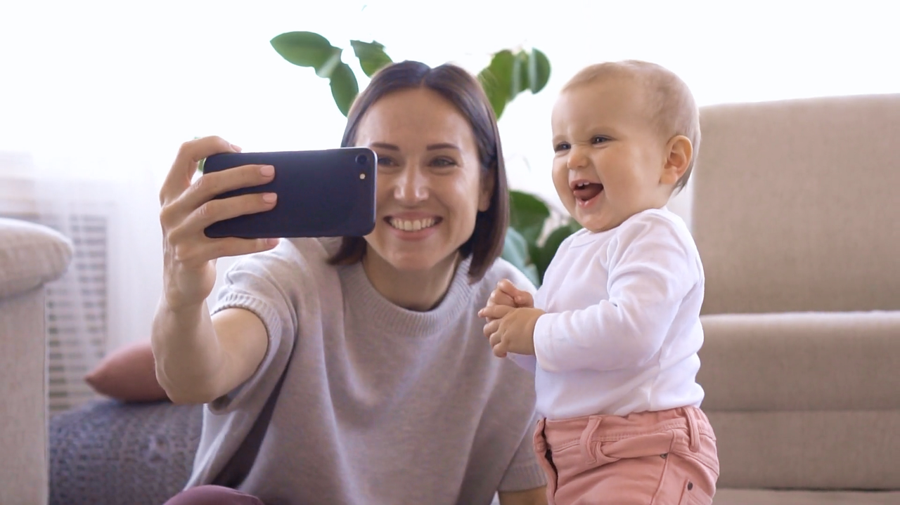 A smiling woman holds a smartphone, taking a selfie with a laughing baby next to her.
