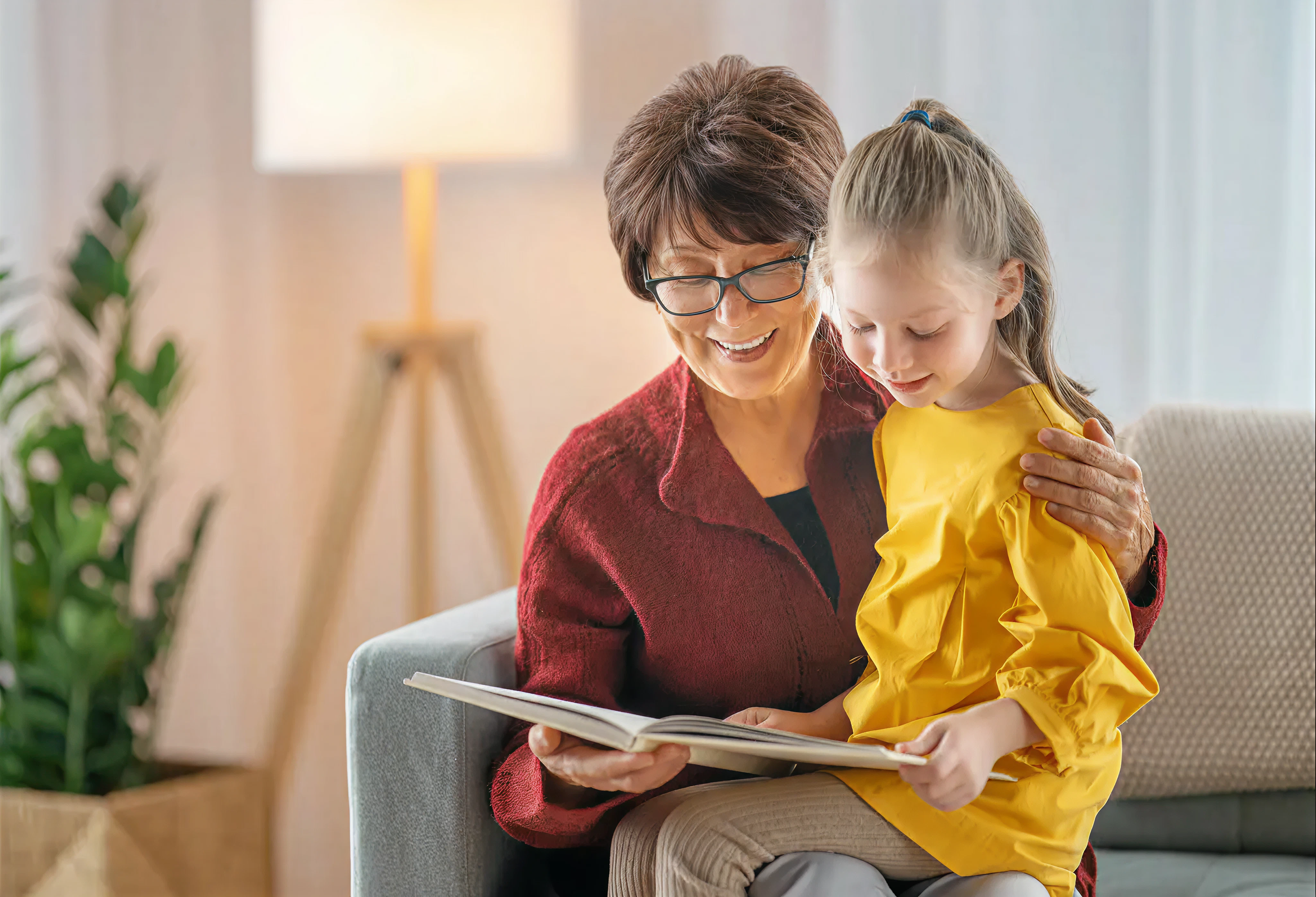 A grandmother sits with a young girl on her lap, and they are reading a book together.