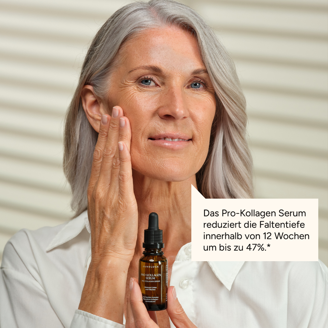 A woman with gray hair holds a bottle of serum while touching her face and smiling.