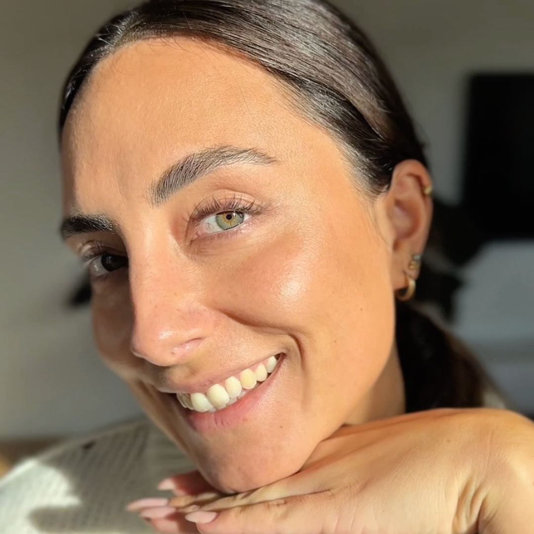 A close-up of a smiling woman with dark hair and light eyes, resting her chin on her hands.