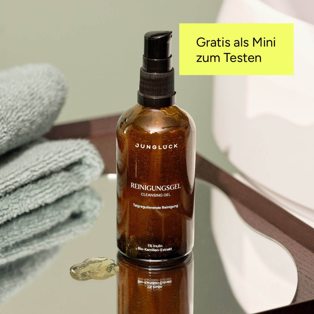 A brown glass pump bottle of Junglück Cleansing Gel sits on a mirrored tray next to a towel.