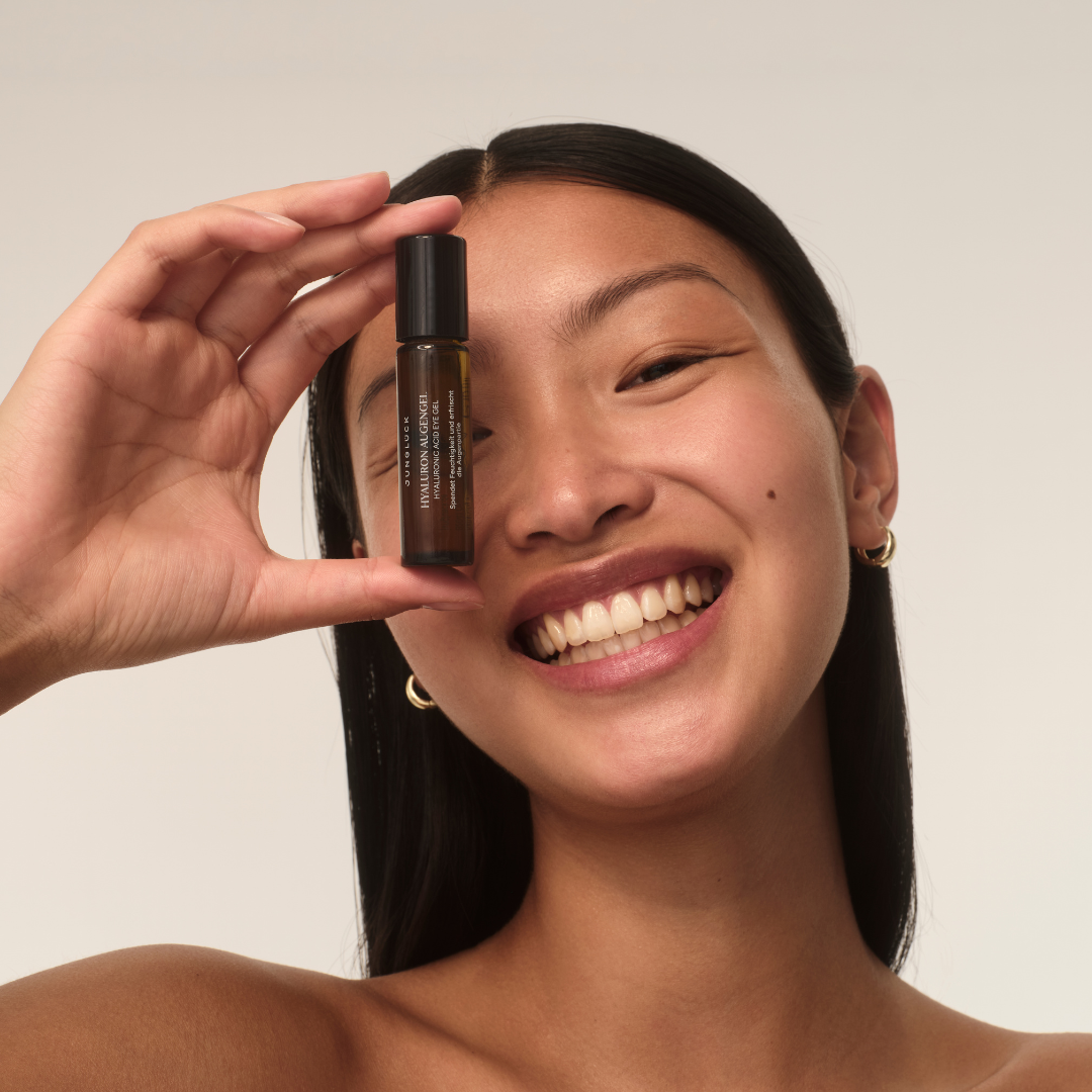 A smiling woman holds a small brown rollerball bottle in front of her eye against a plain background.