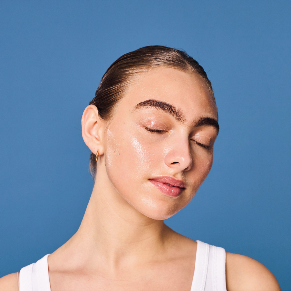 A woman with slicked-back hair and dewy skin poses with her eyes closed against a blue background.