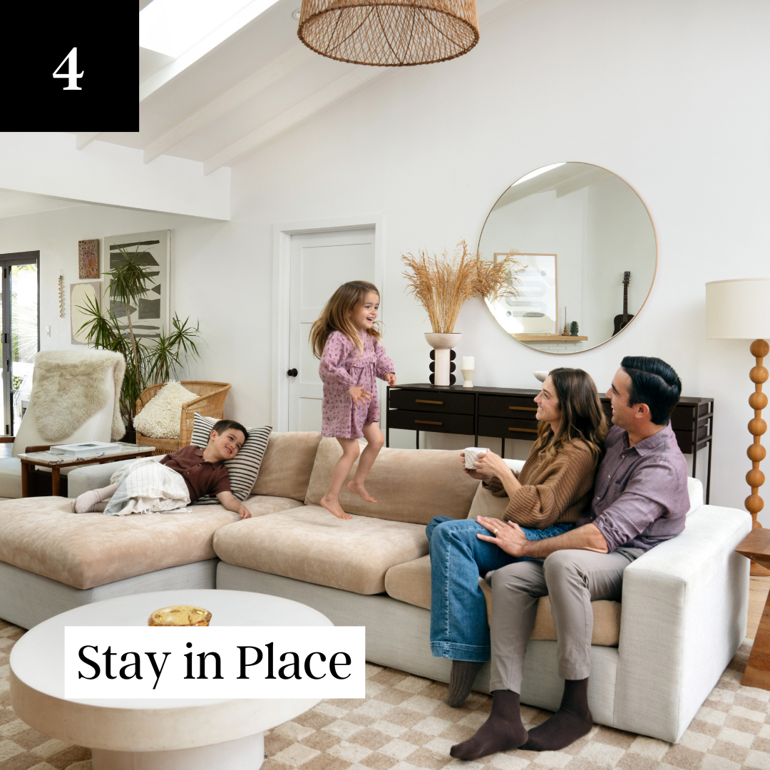 A family of four relaxes in a living room as a young girl jumps on the couch.